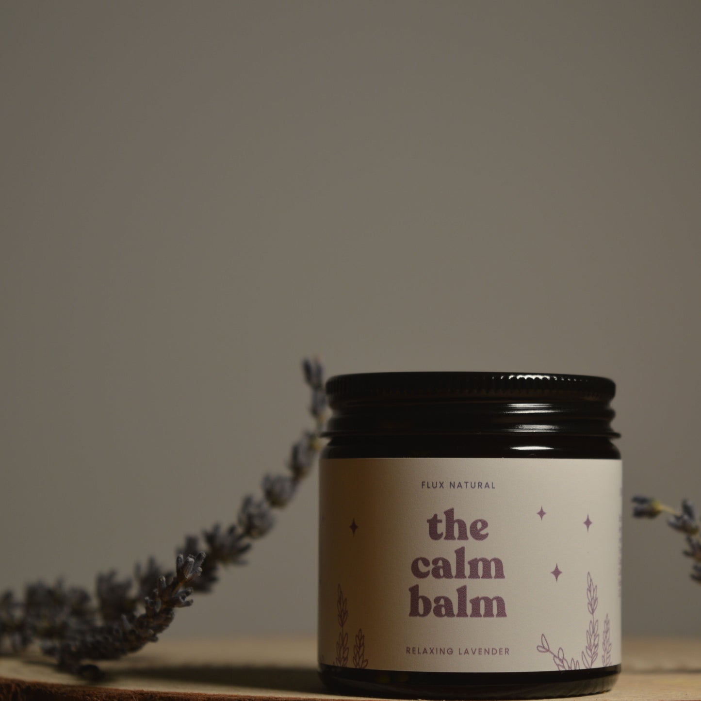 Jar of 'the calm balm' with lavender flowers on a wooden surface and gray background