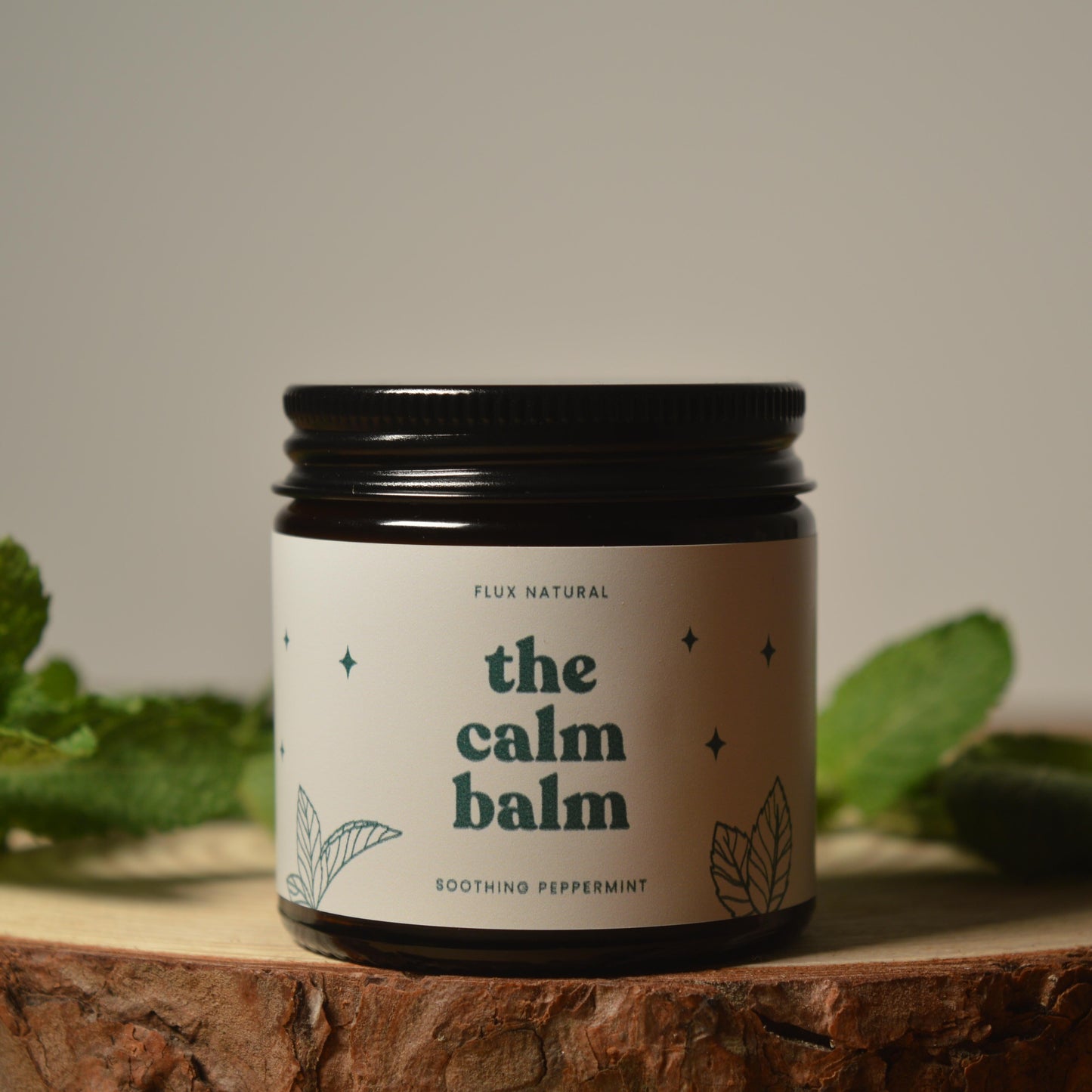 Jar of 'the calm balm' surrounded by peppermint leaves on a wooden surface.