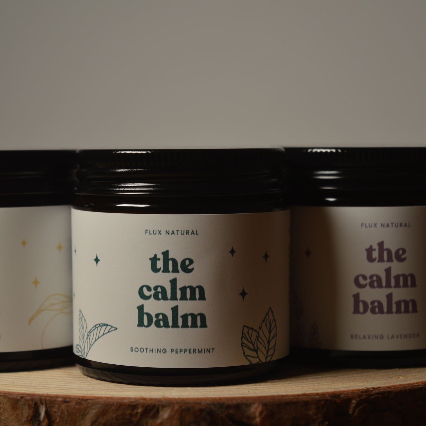Three jars of 'the calm balm' on a wooden surface with a dark background