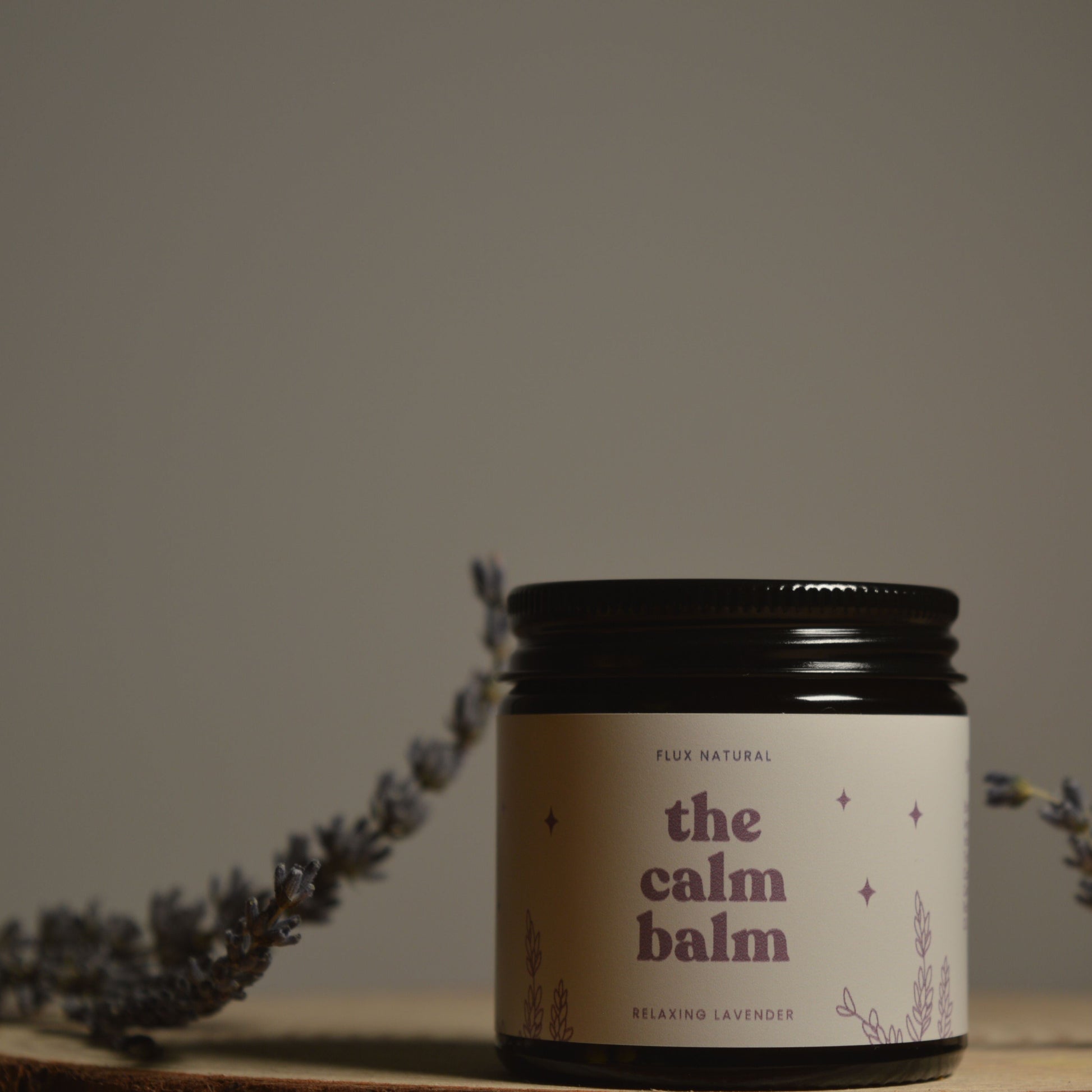 Jar of 'the calm balm' with lavender flowers on a wooden surface and gray background