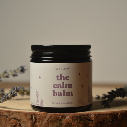 Jar of 'the calm balm' surrounded by lavender flowers on a wooden surface.
