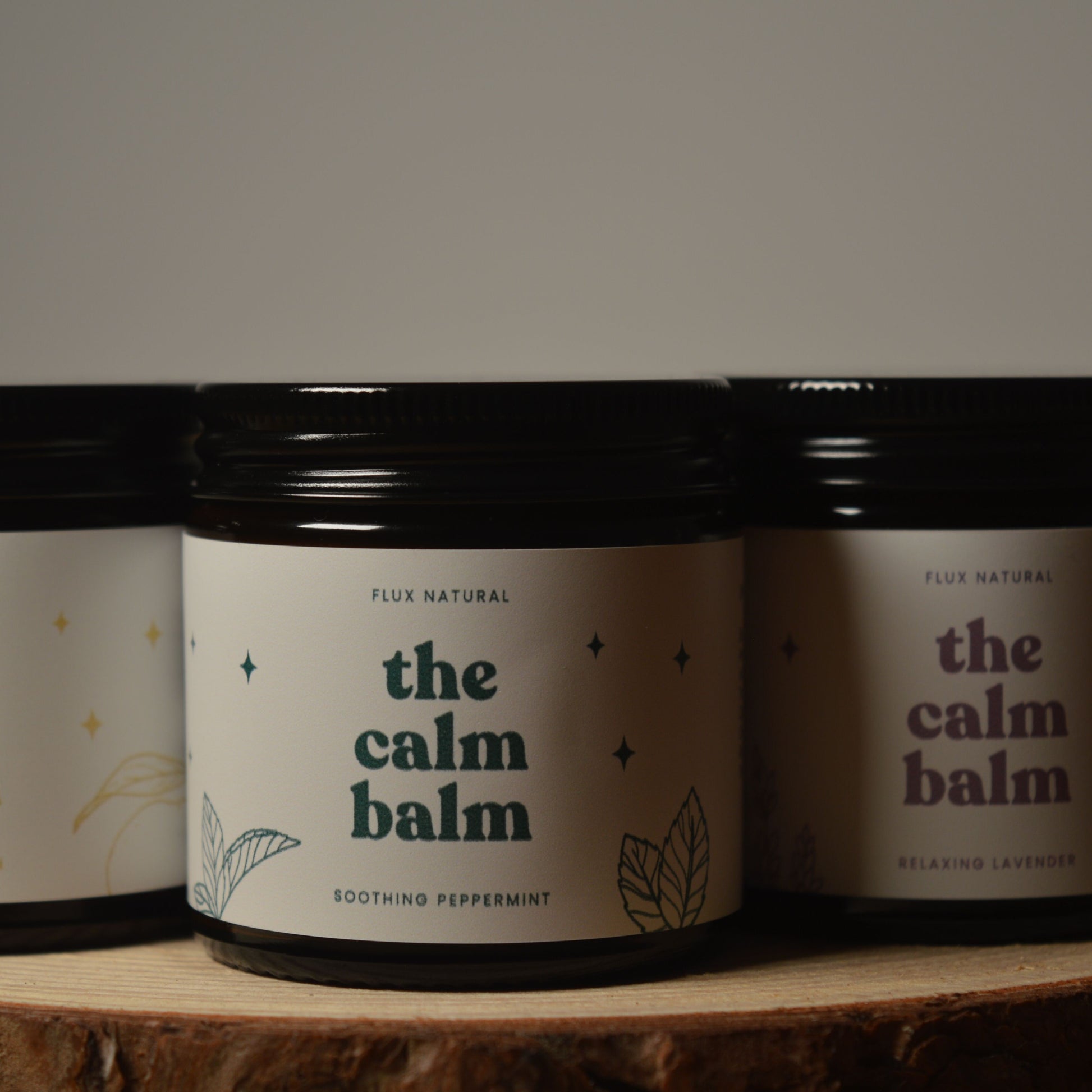 Three jars of 'the calm balm' on a wooden surface with a dark background